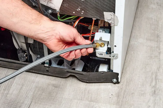 Hand holding the braided water supply line connection behind a refrigerator with an ice maker leaking water