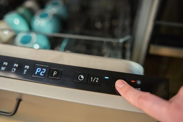 Finger pressing a button beside the illuminated display on a dishwasher's top-edge control panel