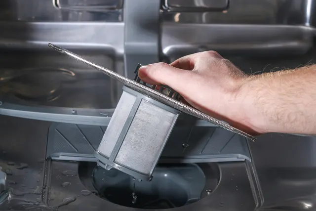 Hand lifting a cylindrical mesh filter from the bottom of a stainless steel dishwasher