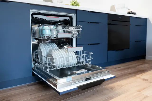 Open dishwasher with racks of plates and cups pulled out in a blue kitchen